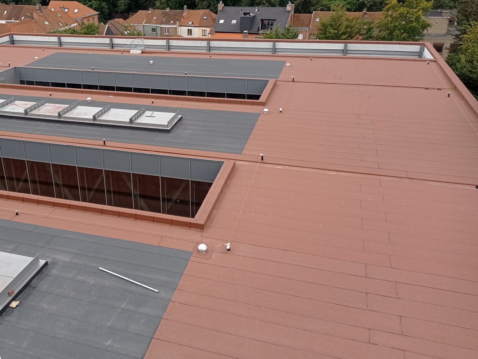 Płaski dach budynku pokryty brązową papą termozgrzewalną z szarymi elementami wokół świetlików, widok z góry na tle zabudowy mieszkalnej i drzew.