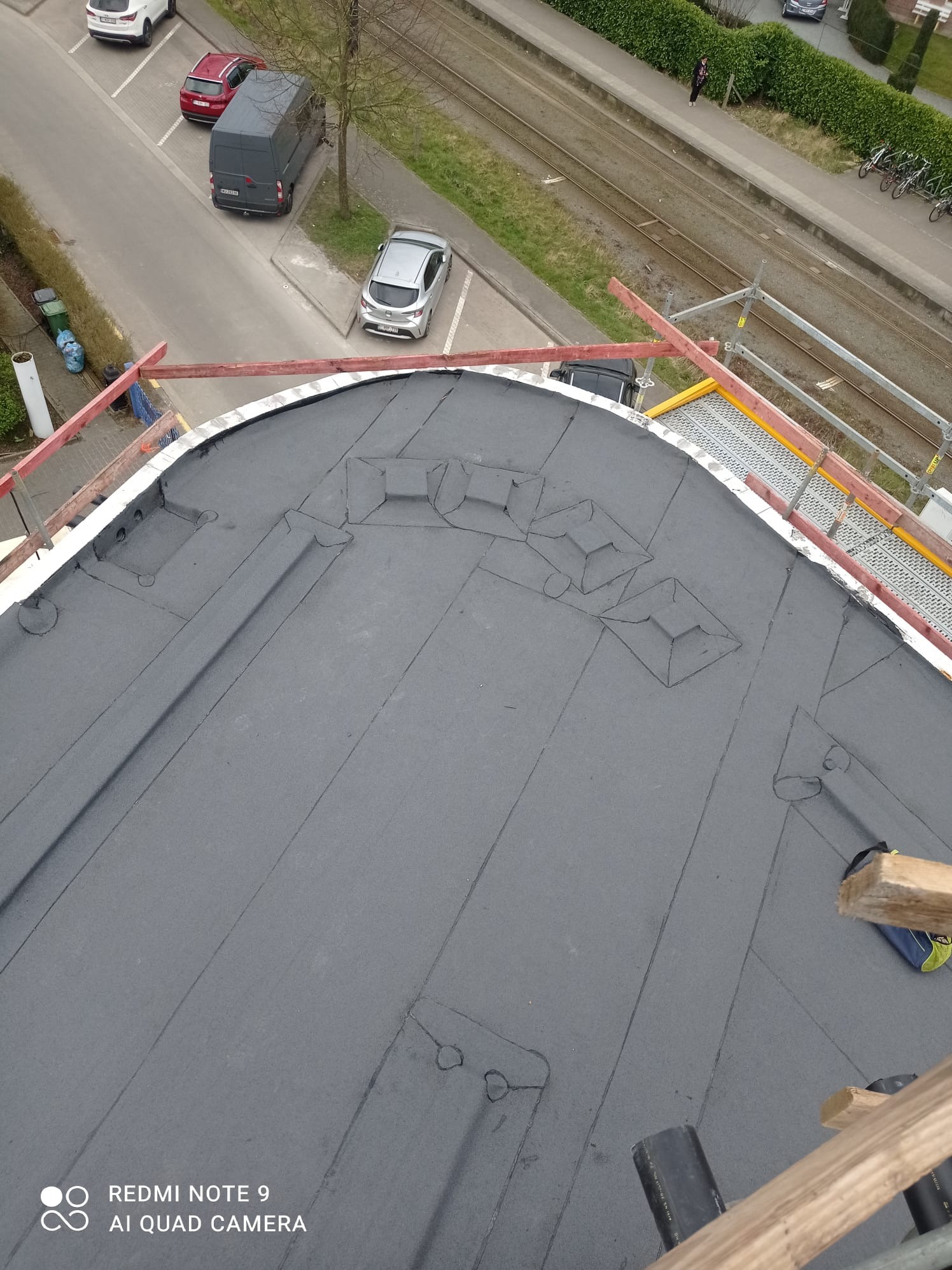 Widok z góry na fragment dachu pokrytego czarną papą, widoczne elementy obróbki blacharskiej i rusztowanie, w tle ulica z zaparkowanymi samochodami i torami kolejowymi.