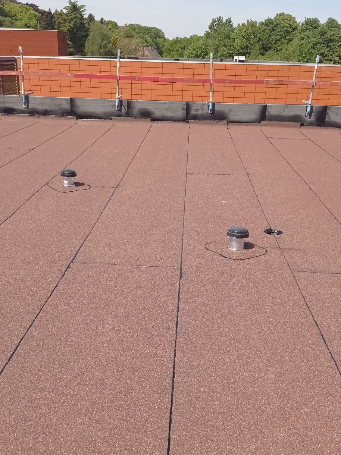 Widok z góry na płaski dach pokryty papą w kolorze ceglastym, z widocznymi dwoma odpowietrznikami i elementami zabezpieczeń na krawędzi dachu.