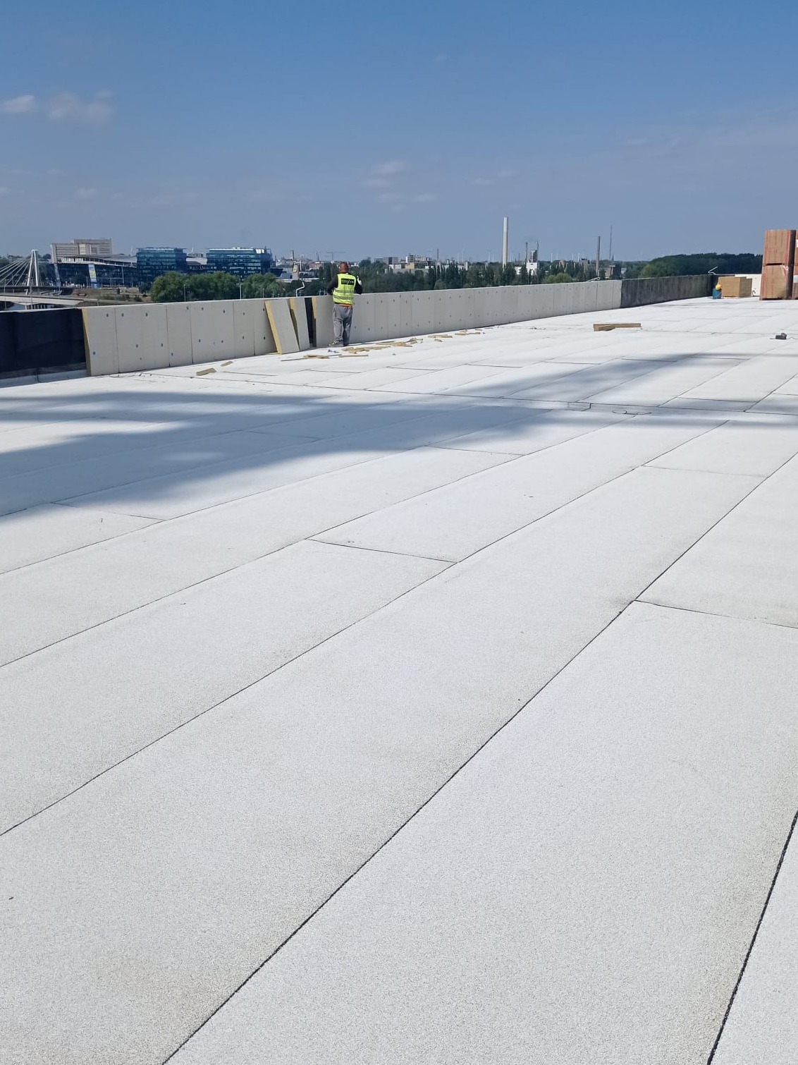 Pracownik w kamizelce odblaskowej na rozległym, płaskim dachu pokrytym jasną membraną, z widocznymi płytami izolacyjnymi i elementami konstrukcyjnymi, na tle panoramy miasta w oddali.