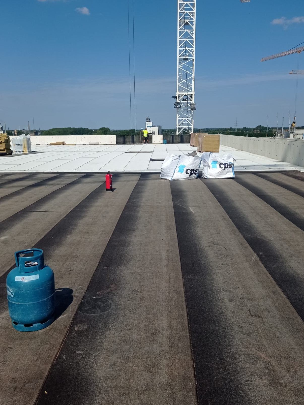 Widok z góry na płaski dach w trakcie prac, z widocznymi pasami papy termozgrzewalnej, butlą gazową, gaśnicą, workami z materiałem izolacyjnym oraz pracownikiem w oddali i żurawiem w tle.