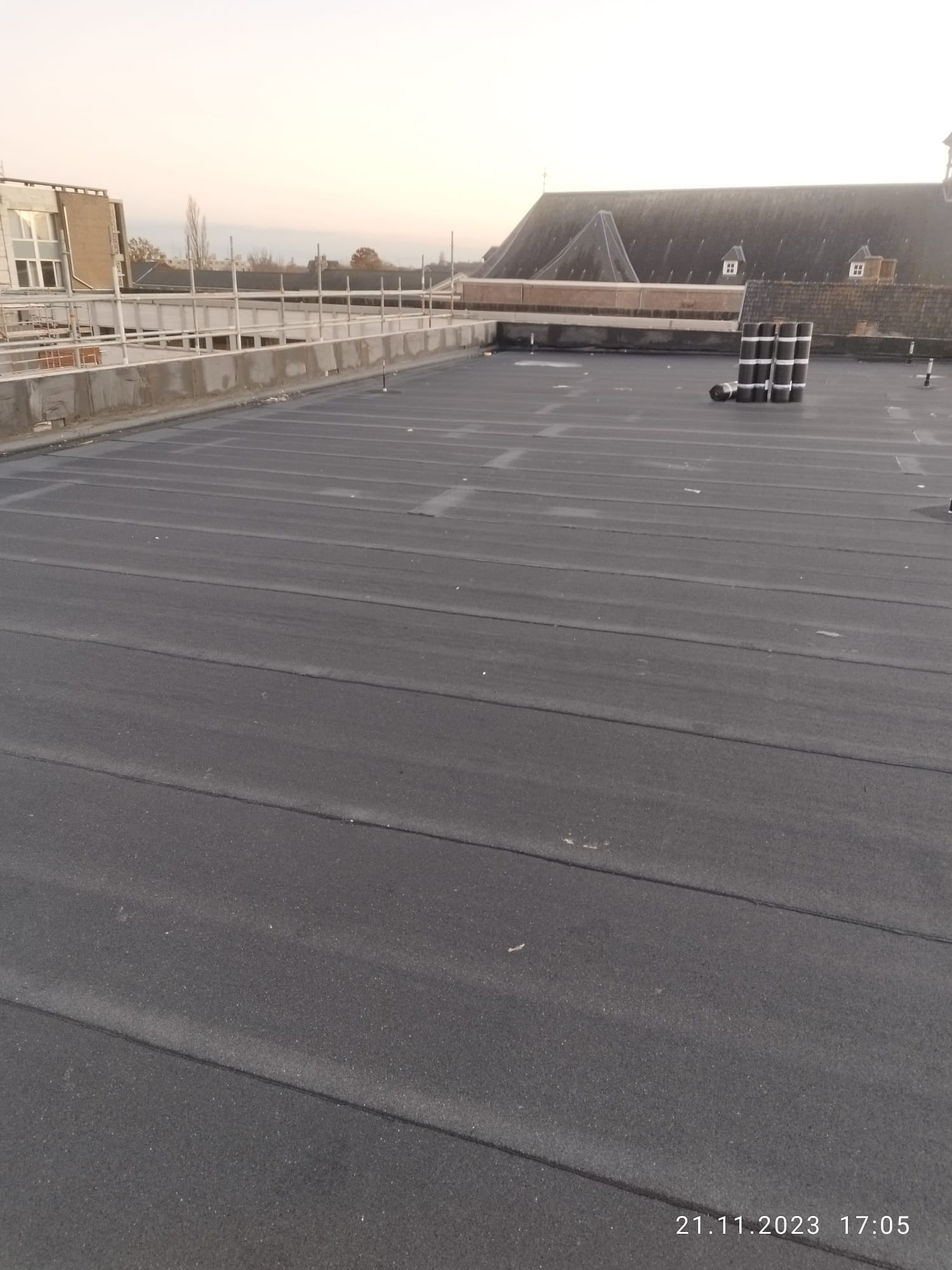 Widok z góry na płaski dach pokryty papą termozgrzewalną, z widocznymi rolkami papy i rusztowaniem w tle, w pochmurny dzień.