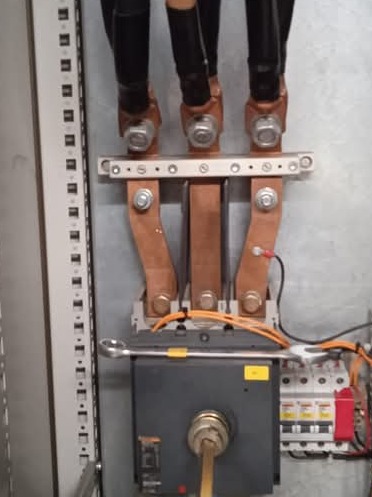 Szyna zasilająca z miedzianymi zaciskami i podłączonymi kablami, obok wyłącznik z kluczem i listwa bezpieczników, klucz płaski leży na wyłączniku.