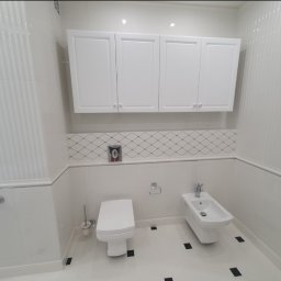 WP-BUD - Jasna łazienka z białymi szafkami, toaletą i bidetem. Na podłodze białe płytki z czarnymi akcentami. Ściany wykończone w białej tonacji z delikatnym wzorem.