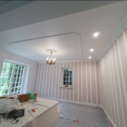 WP-BUD - Pokój po odświeżeniu: biały sufit z dekoracyjną sztukaterią i oświetleniem punktowym, ściany w beżowo-białe pasy, okna z widokiem na zieleń. Częściowo zakryte meble.