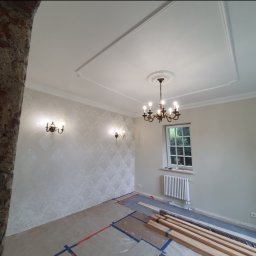 WP-BUD - Pokój po remoncie: wzorzysta tapeta, ozdobne listwy sufitowe, żyrandol. Na podłodze zabezpieczenia i drewniane elementy. Widoczny fragment ceglanej ściany.