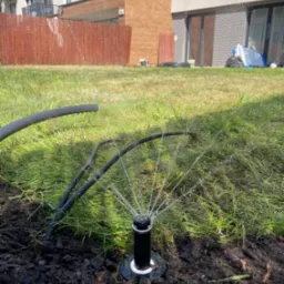 Działający zraszacz turbinowy nawadnia trawnik przed budynkiem mieszkalnym. W tle widoczny płot z wikliny i budynek z balkonami. Zbliżenie na zainstalowany system nawadniania.