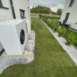 NOIBUD - Ułożona trawa z rolki wzdłuż ścieżki z białego grysu, obok budynku z pompami ciepła. W tle osoba grabiąca trawę. Zadbany ogród.