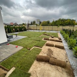 NOIBUD - Układanie trawy z rolki w ogrodzie. Częściowo ułożona darń i zwinięte rolki na przygotowanym podłożu. W tle widoczny dom z elementami małej architektury.