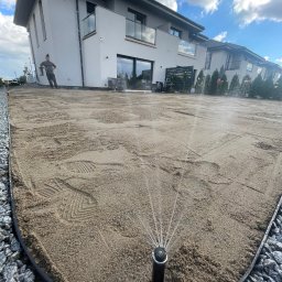 NOIBUD - Automatyczne nawadnianie w trakcie przygotowania terenu pod trawnik, widoczne ślady stóp na piasku i nowoczesny dom w tle, ozdobne kamienie na obrzeżach trawnika.