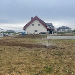 NOIBUD - Rozpoczęte nawadnianie: zraszacze w akcji na przygotowanym terenie pod ogród, w tle dom z czerwoną dachówką i ogrodzenie z siatki. Pochmurne niebo.