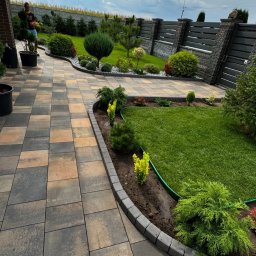 NOIBUD - Ukończony ogród z trawnikiem, ścieżką z kostki brukowej i ozdobnymi nasadzeniami, otoczony nowoczesnym ogrodzeniem. Widoczny fragment systemu nawadniania.