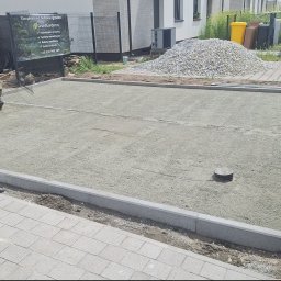 Noibud - Pracownik wyrównuje podłoże z kruszywa, przygotowując teren pod układanie kostki brukowej, otoczone krawężnikami. W tle stos kostki i tablica firmy ogrodniczej.