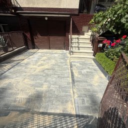 Noibud - Nowo ułożony podjazd z szarej kostki brukowej z resztkami piasku do fugowania, prowadzący do brązowego garażu. Obok betonowe schody, metalowe balustrady i zadbane krzewy z różami.