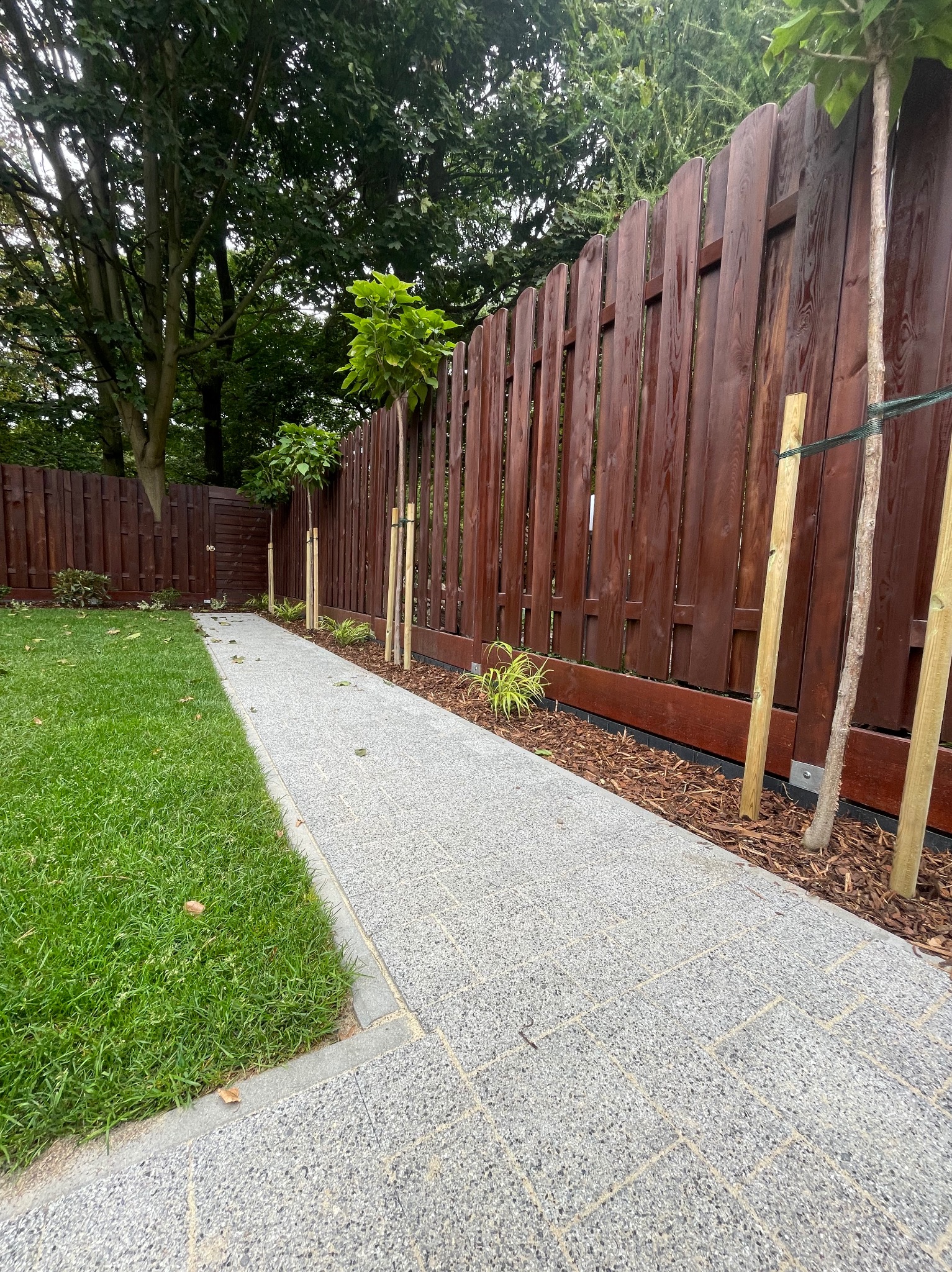 Ścieżka z kostki brukowej w ogrodzie, obsadzona młodymi drzewkami i oddzielona od trawnika drewnianym płotem. Widoczne świeże nasadzenia i kora.