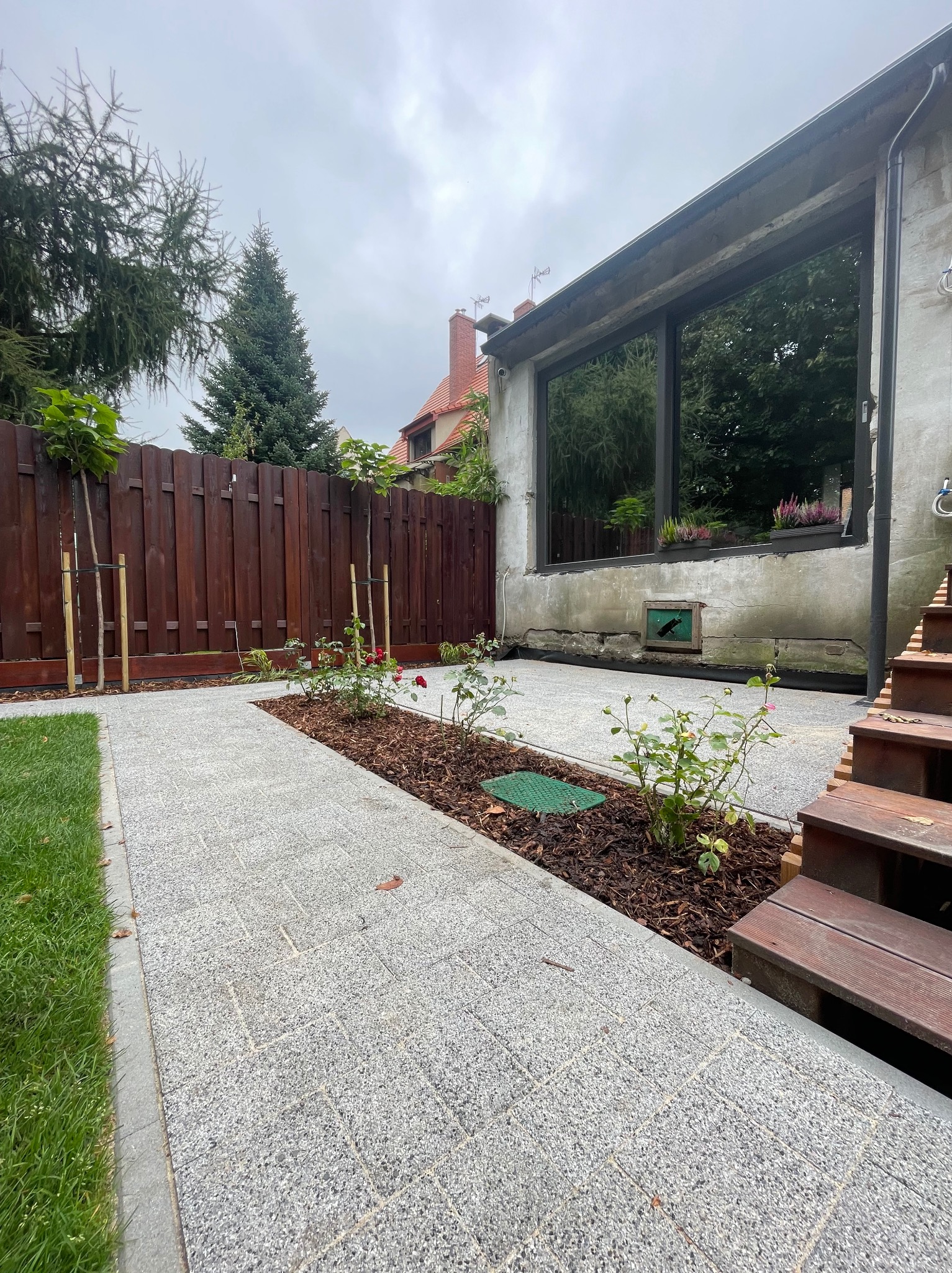 Ścieżka z kostki granitowej w ogrodzie z rabatami róż, drewnianym ogrodzeniem i schodami. Widoczne drzewa i budynek w tle.