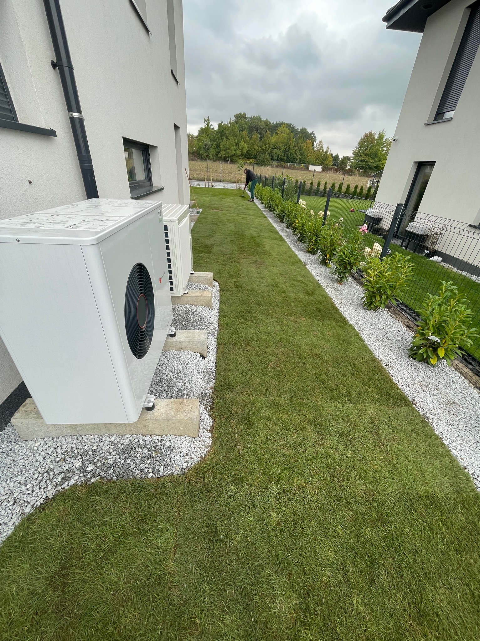 Ułożona trawa z rolki wzdłuż ścieżki z białego grysu, obok budynku z pompami ciepła. W tle osoba grabiąca trawę. Zadbany ogród.