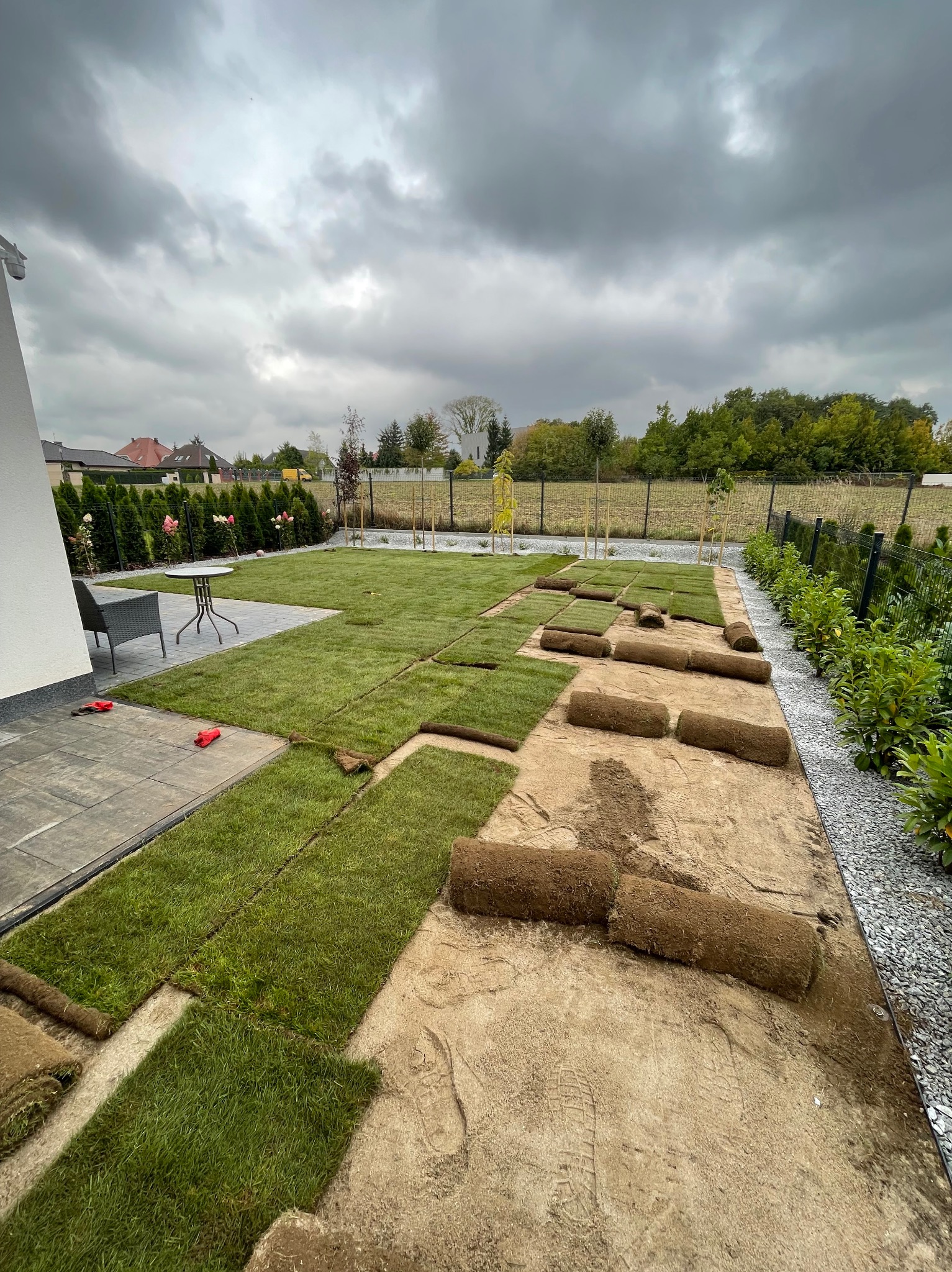 Układanie trawy z rolki w ogrodzie. Częściowo ułożona darń i zwinięte rolki na przygotowanym podłożu. W tle widoczny dom z elementami małej architektury.
