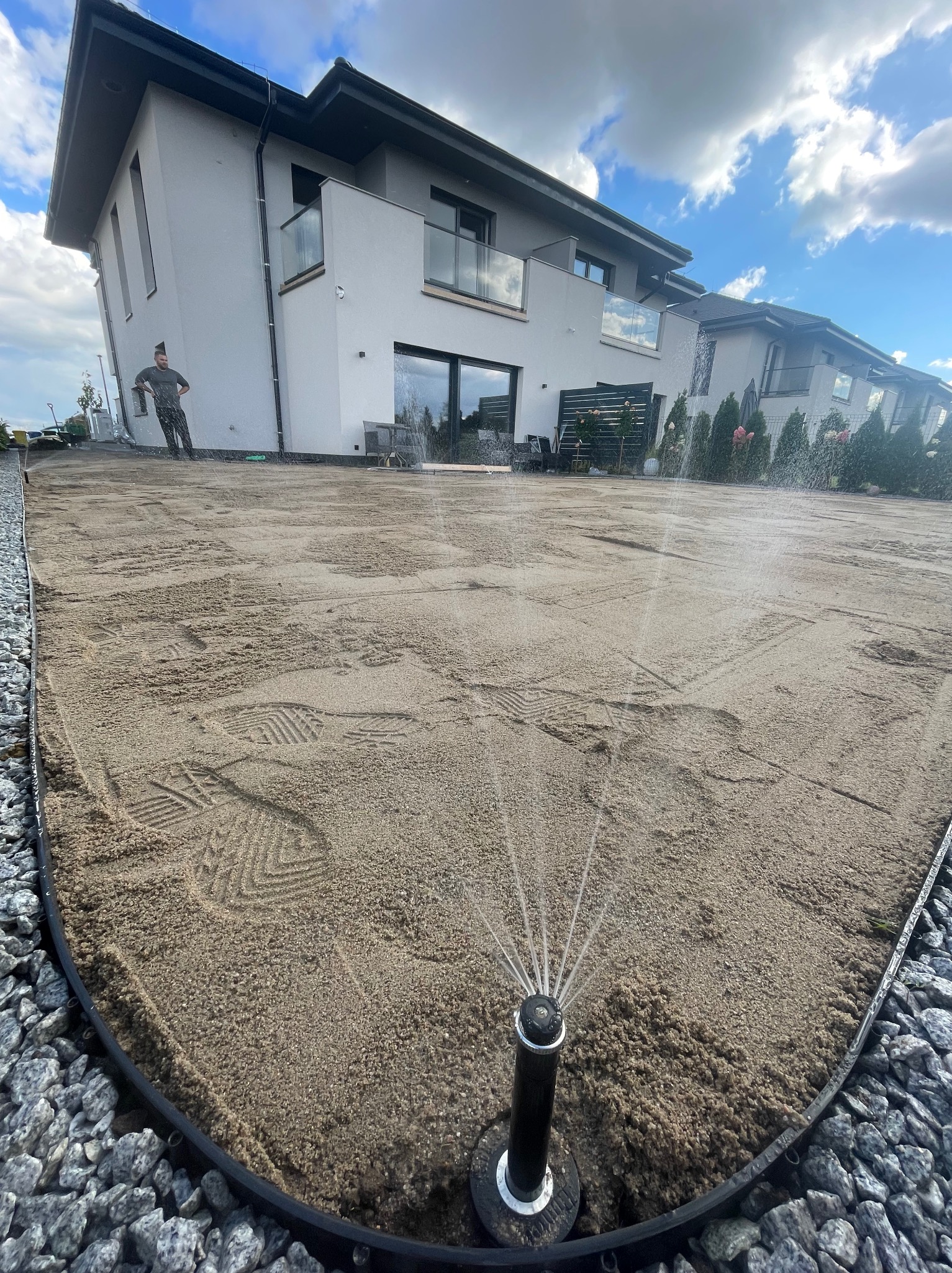 Automatyczne nawadnianie w trakcie przygotowania terenu pod trawnik, widoczne ślady stóp na piasku i nowoczesny dom w tle, ozdobne kamienie na obrzeżach trawnika.