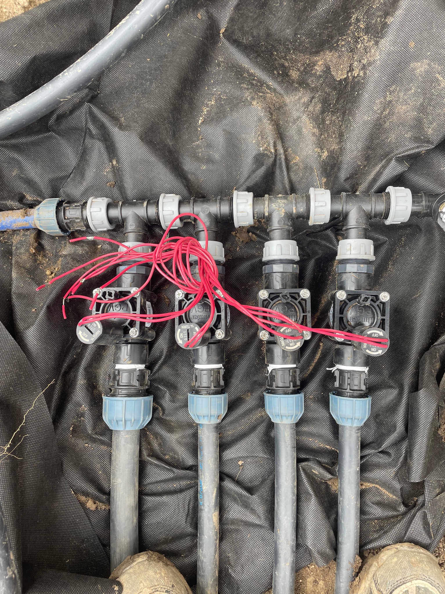 Fragment instalacji systemu nawadniania z zaworami i przewodami elektrycznymi na czarnej macie w ziemi. Prace ogrodnicze we Wrocławiu.