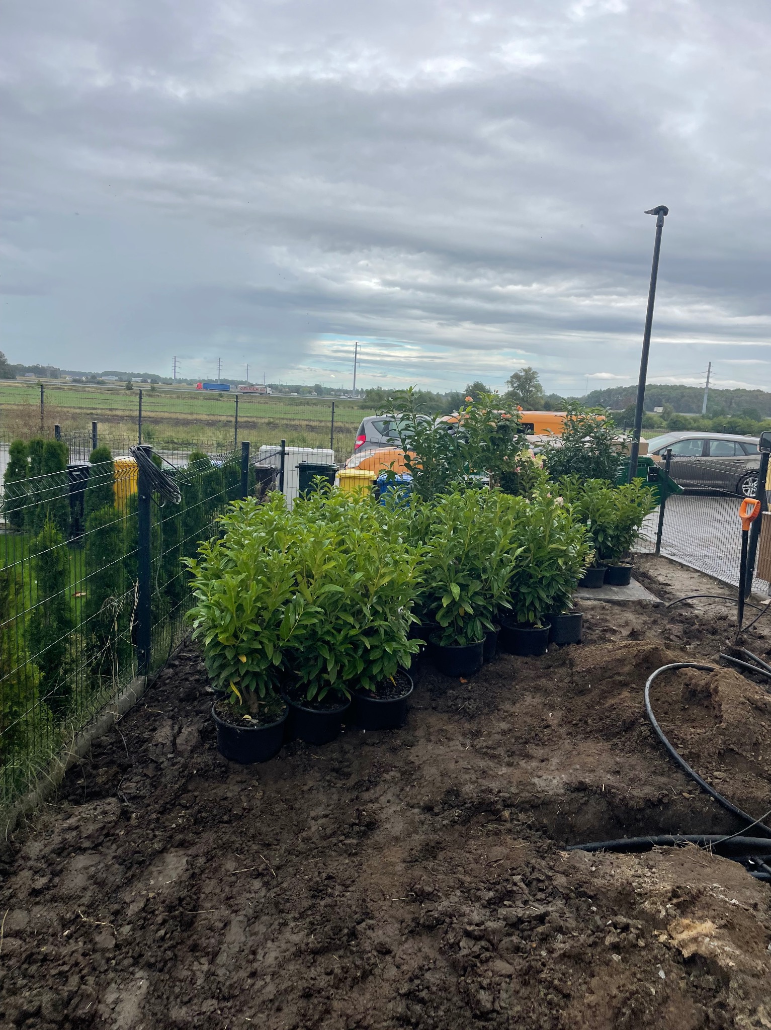 Przygotowanie do sadzenia roślin w donicach wzdłuż ogrodzenia. Widoczne narzędzia i elementy instalacji nawadniającej. Pochmurne niebo w tle.