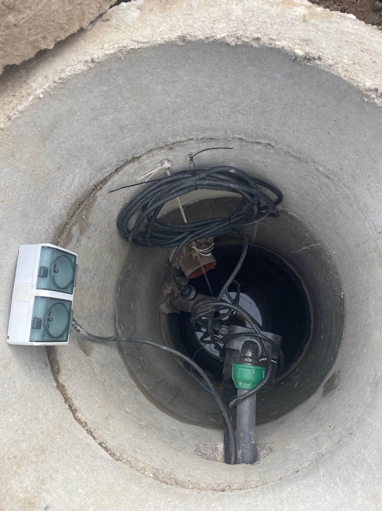 Studnia betonowa z pompą głębinową i kablem zasilającym. Element systemu nawadniania ogrodu. Widok z góry na wnętrze studni z instalacją.