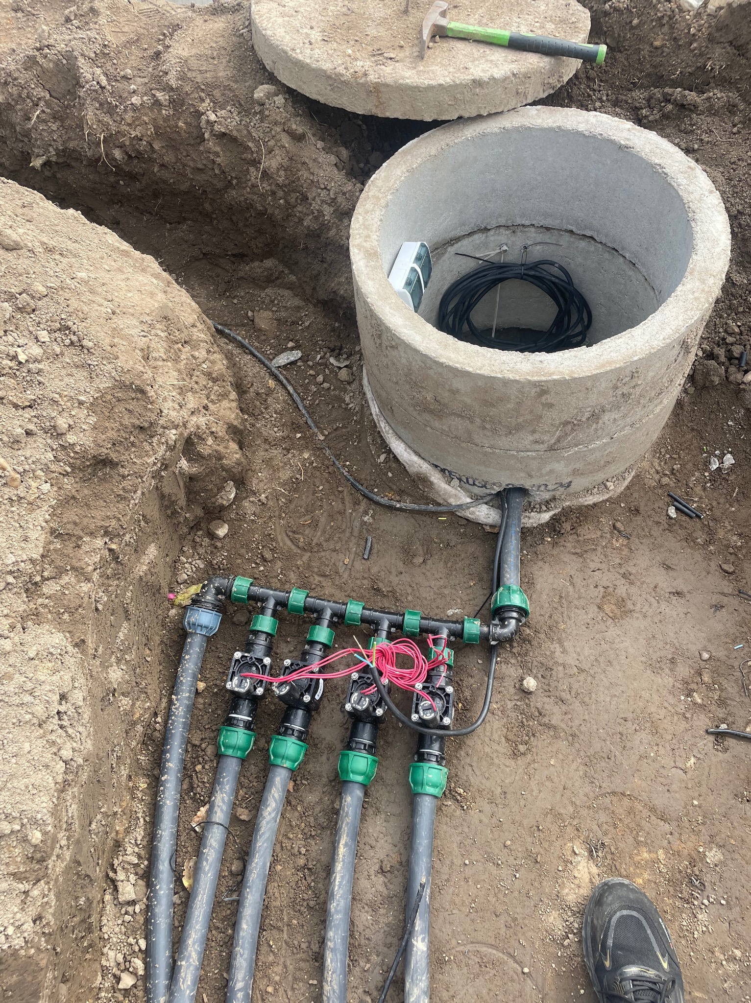 Montaż systemu nawadniania: betonowy krąg studni z przewodami, rozdzielacz z zaworami i rury w wykopie. Widoczny młotek i but. Detal instalacji w ziemi.