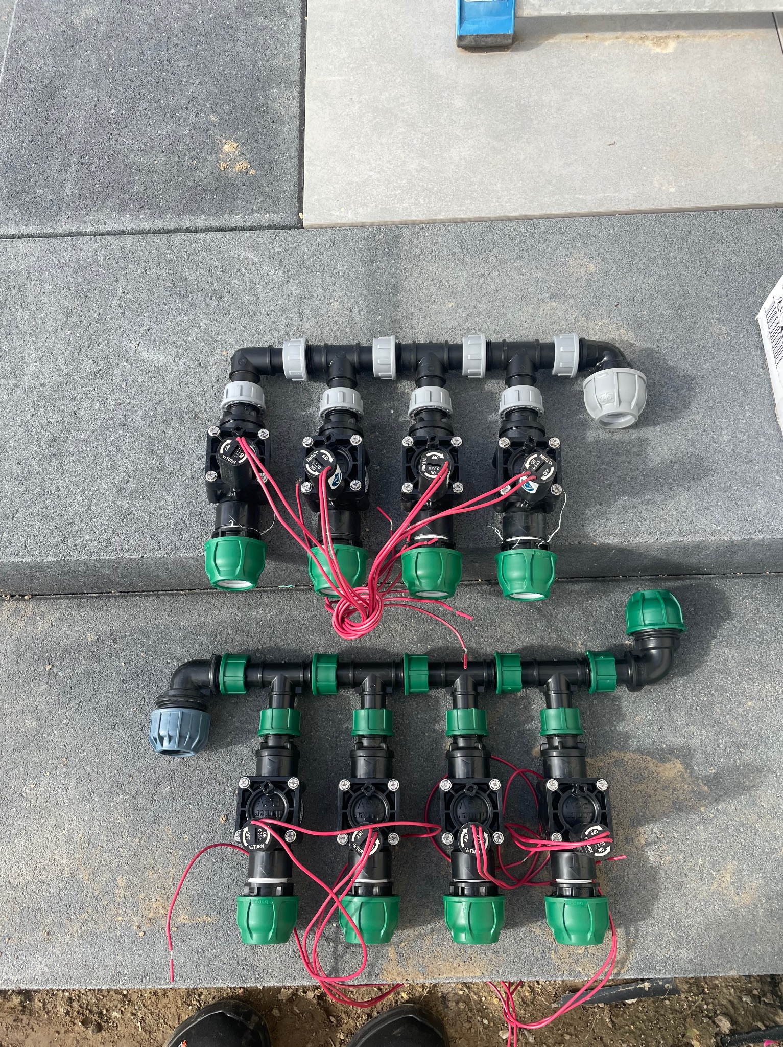 Rozłożony system nawadniania z zaworami elektromagnetycznymi na szarych płytach chodnikowych. Widoczne złącza, rury i przewody elektryczne.
