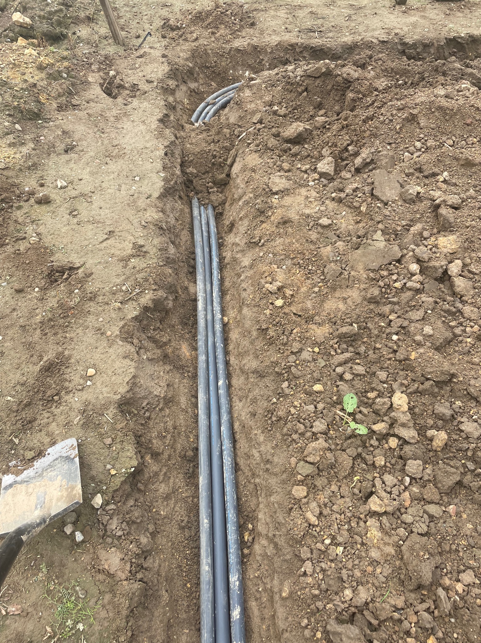 Wkopane rury w ziemi, prawdopodobnie system nawadniania. Widoczny fragment łopaty. Ujęcie z góry, ziemia z kamieniami.