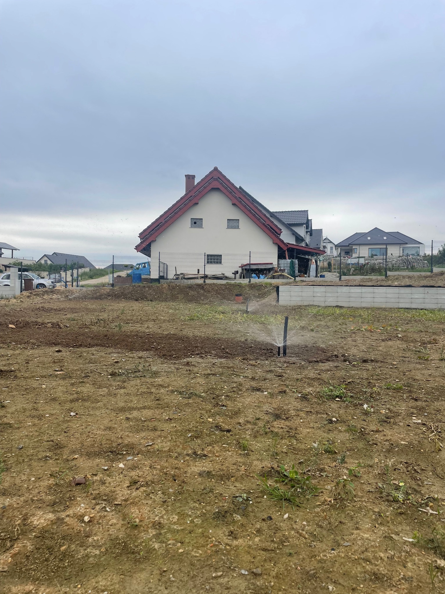 Rozpoczęte nawadnianie: zraszacze w akcji na przygotowanym terenie pod ogród, w tle dom z czerwoną dachówką i ogrodzenie z siatki. Pochmurne niebo.