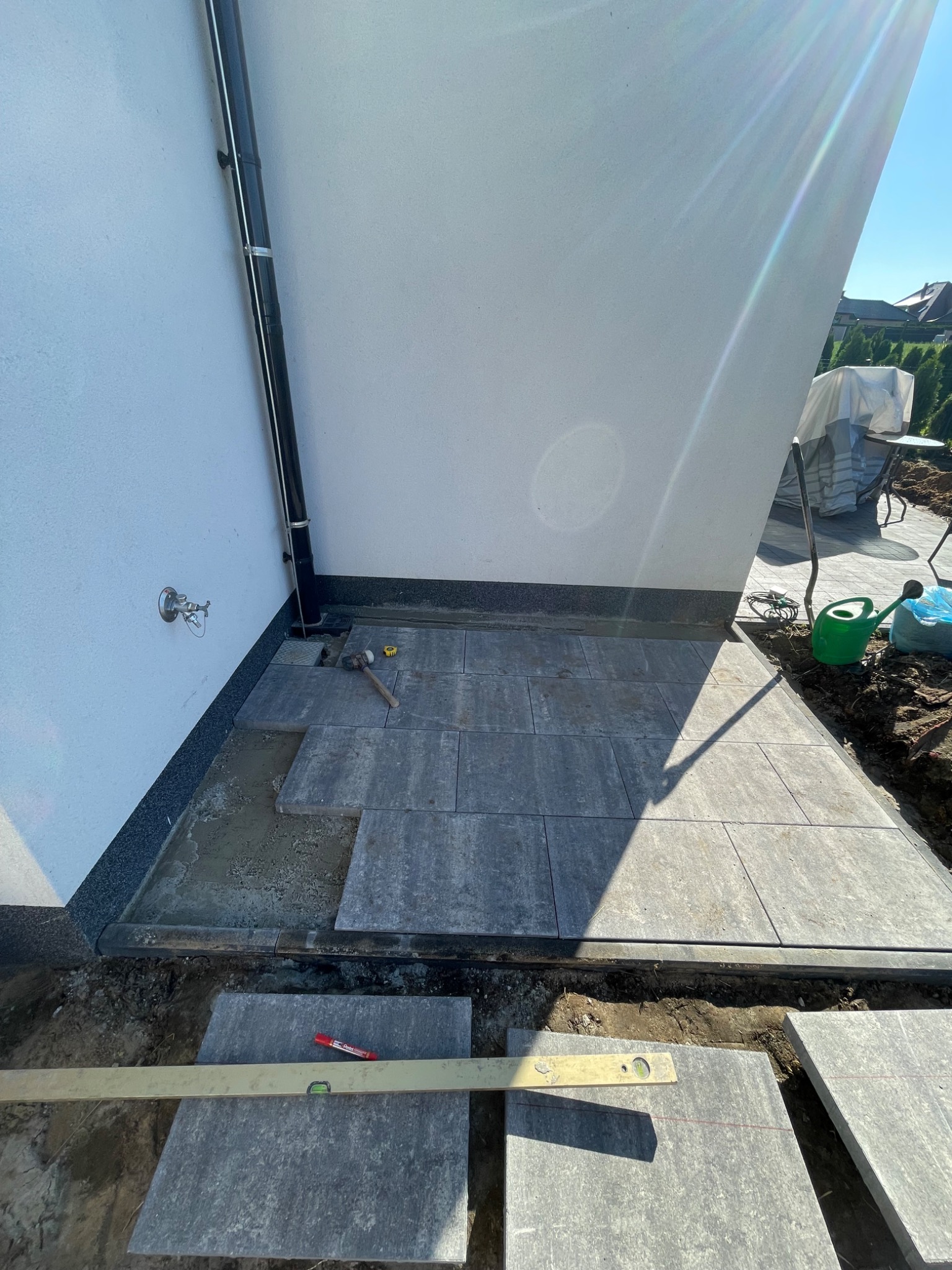 Układanie szarej kostki brukowej na tarasie przy ścianie budynku. Widoczny poziomica, gumowy młotek i zielona konewka. Częściowo ułożona nawierzchnia.