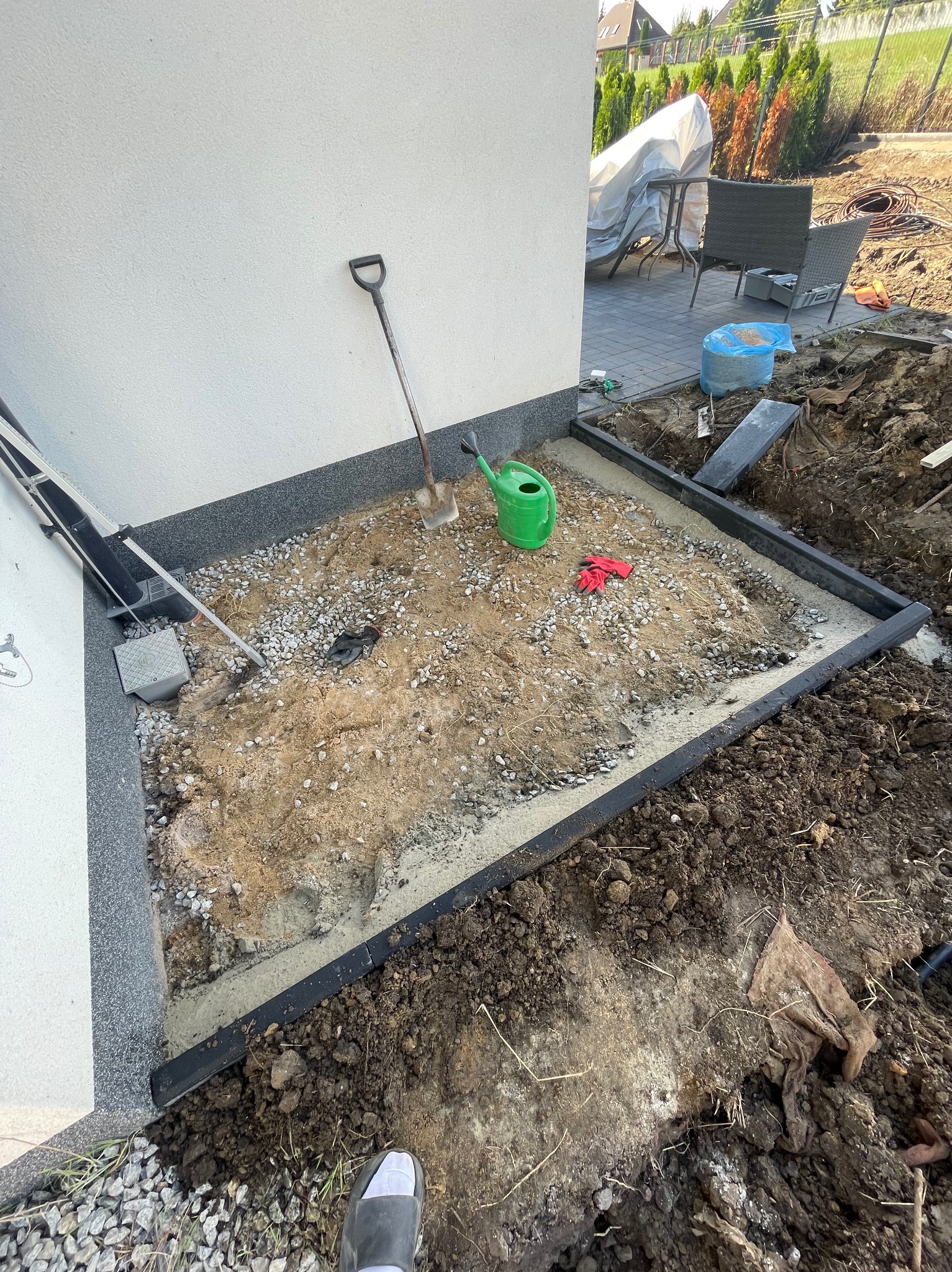 Przygotowany teren pod trawnik: obrzeża z kostki, podsypka, narzędzia ogrodnicze (szpadel, konewka), rękawice robocze. Widok z góry na plac budowy.