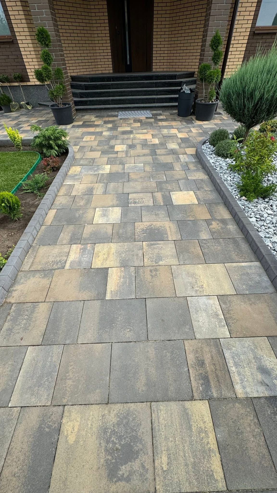 Ułożona kostka brukowa w odcieniach szarości i beżu tworzy ścieżkę prowadzącą do domu. Po bokach rabaty z roślinami i kamieniami dekoracyjnymi.