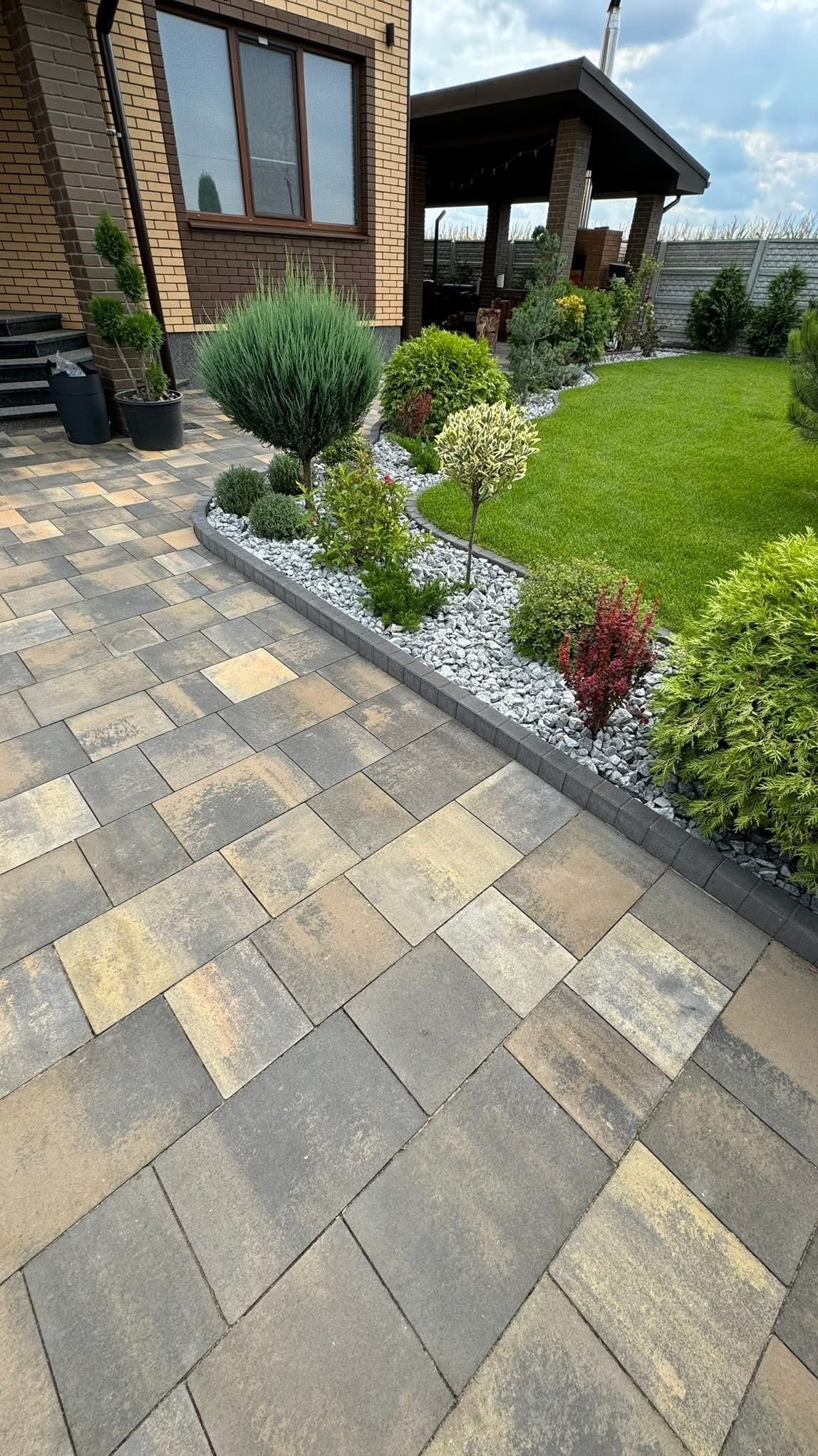 Ułożona kostka brukowa w odcieniach szarości i beżu, obrzeża z kamienia, rabata z roślinami ozdobnymi i trawnik na tle domu z cegły i altany.