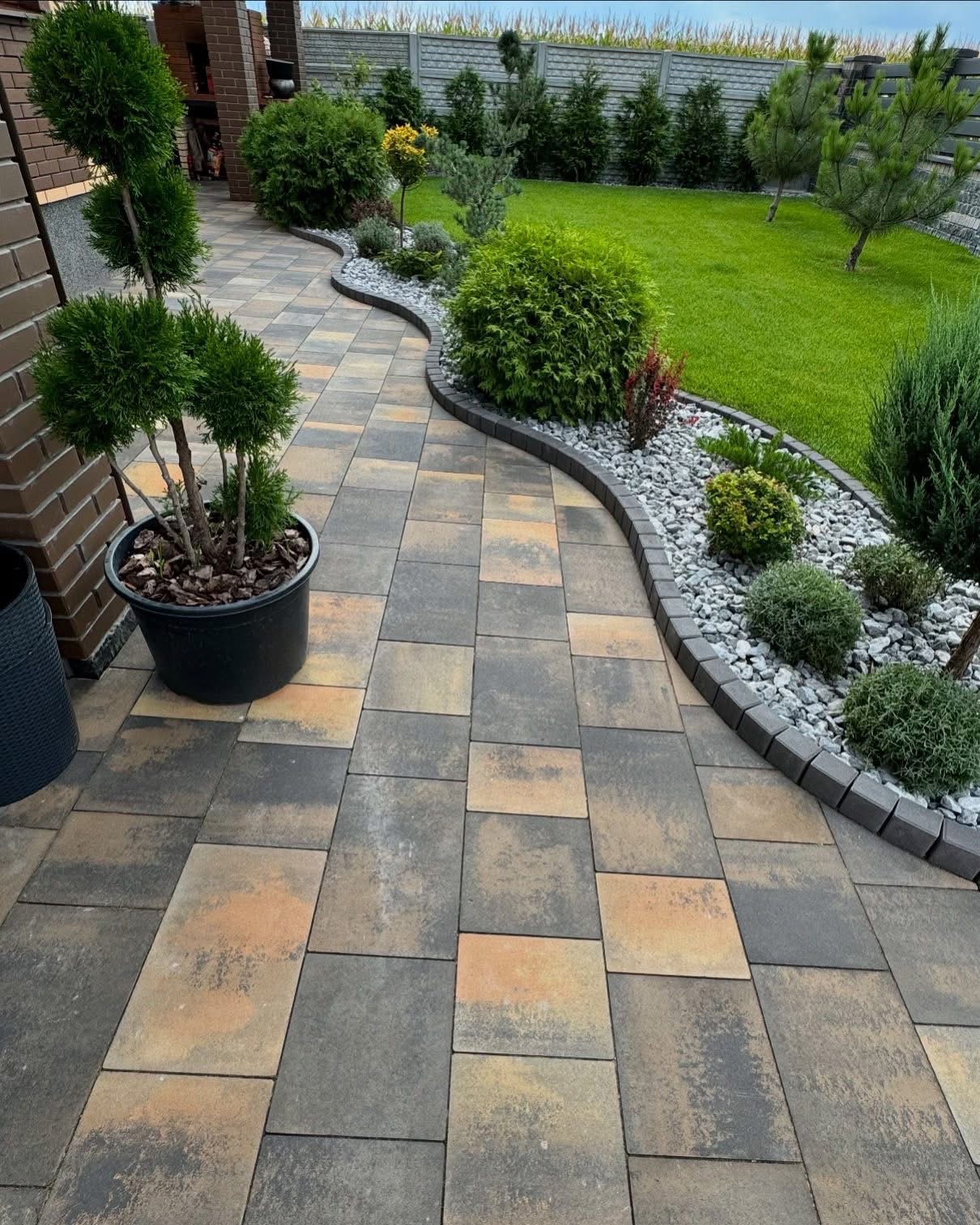 Kaskadowa ścieżka z kostki brukowej prowadzi przez zadbany ogród z trawnikiem, ozdobnymi krzewami i drzewami. Donica z formowanym drzewkiem przy budynku.