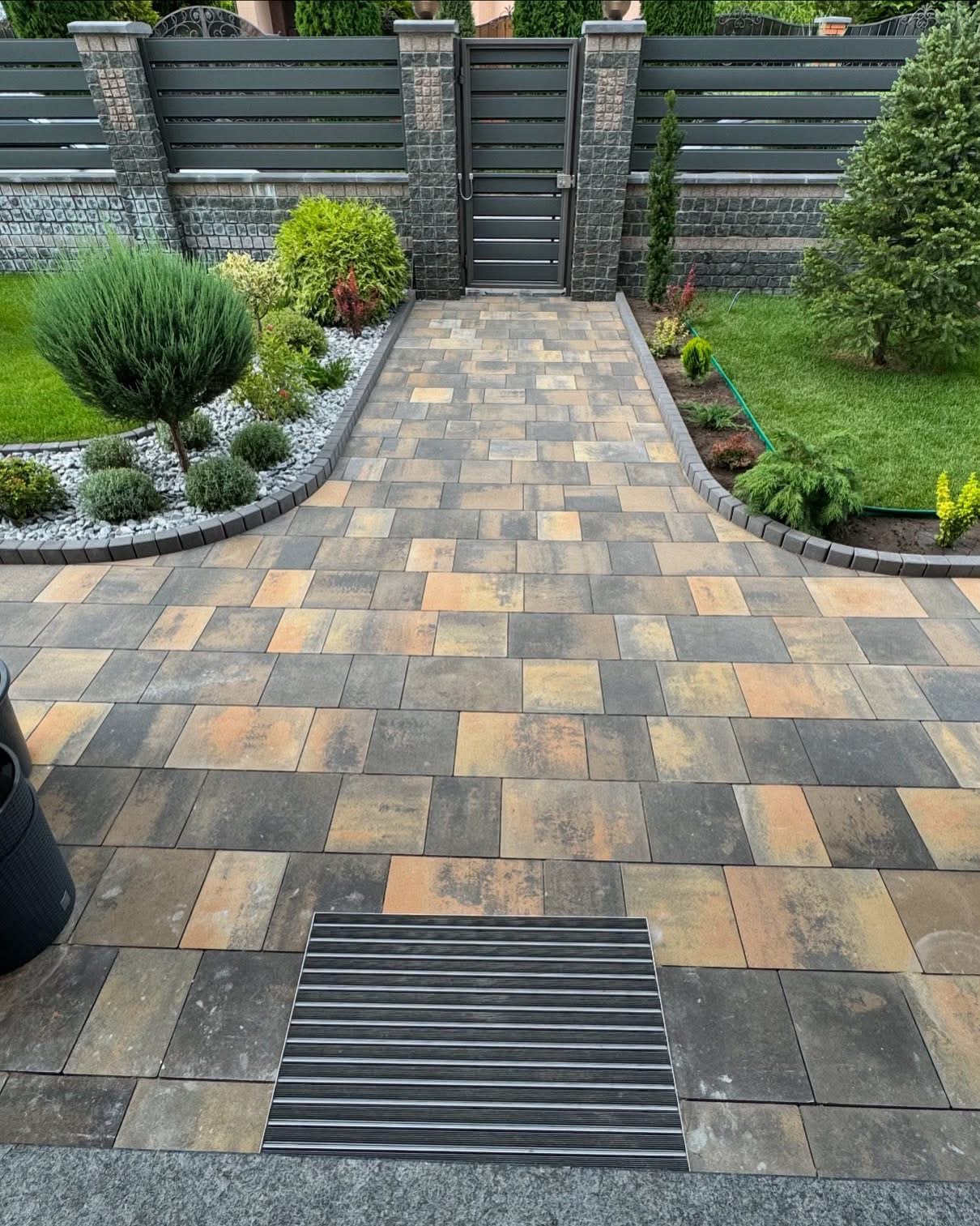 Ułożona kostka brukowa w ogrodzie prowadząca do furtki. Po bokach rabaty z roślinami i kamieniami. Widoczny fragment trawnika i ogrodzenia.