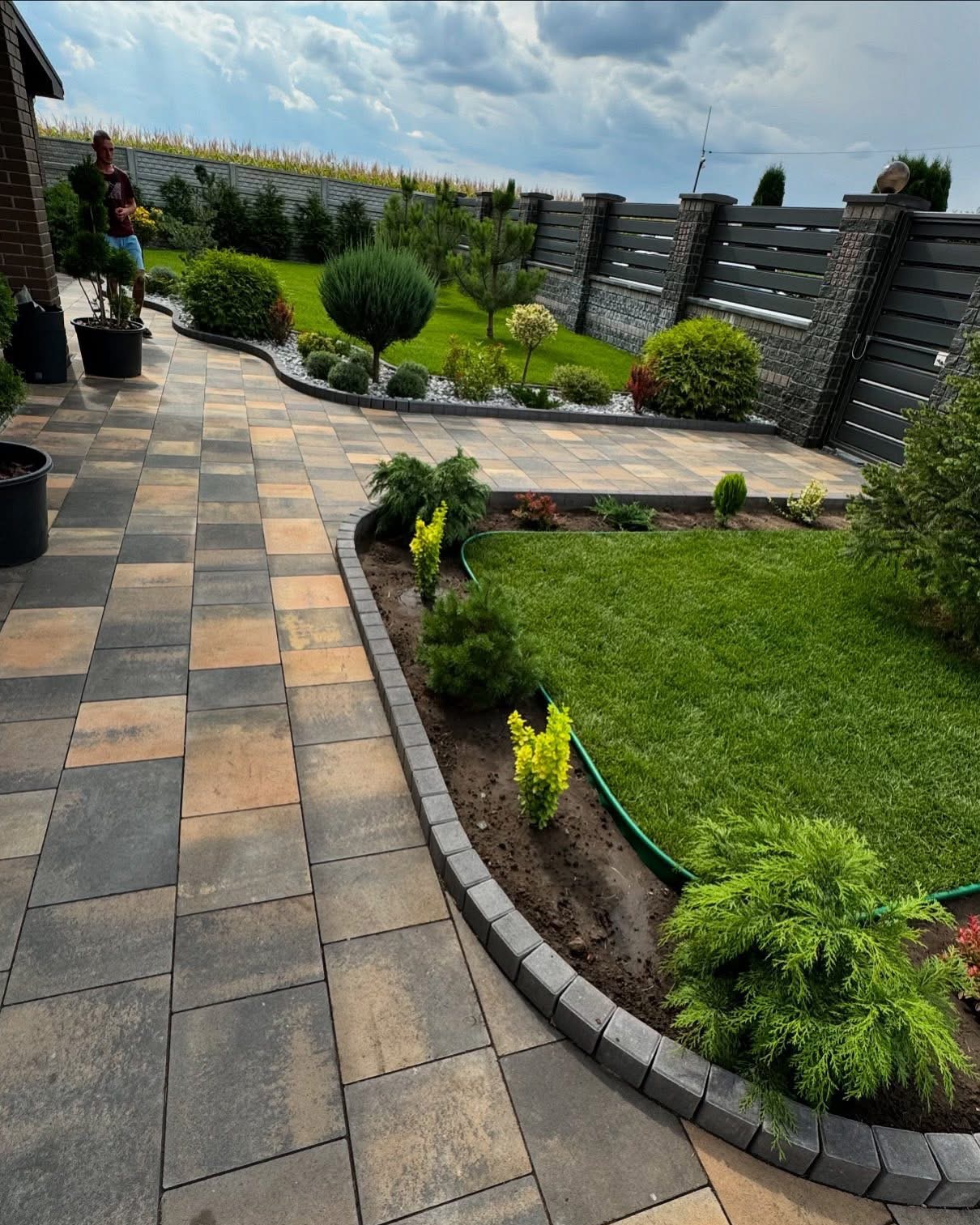 Ukończony ogród z trawnikiem, ścieżką z kostki brukowej i ozdobnymi nasadzeniami, otoczony nowoczesnym ogrodzeniem. Widoczny fragment systemu nawadniania.