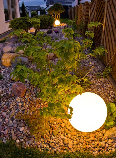 Oświetlony ogród z kulistymi lampami, kamieniami i roślinami. Dekoracyjne oświetlenie podkreśla aranżację ogrodu po zmroku. Widoczny fragment drewnianego ogrodzenia.