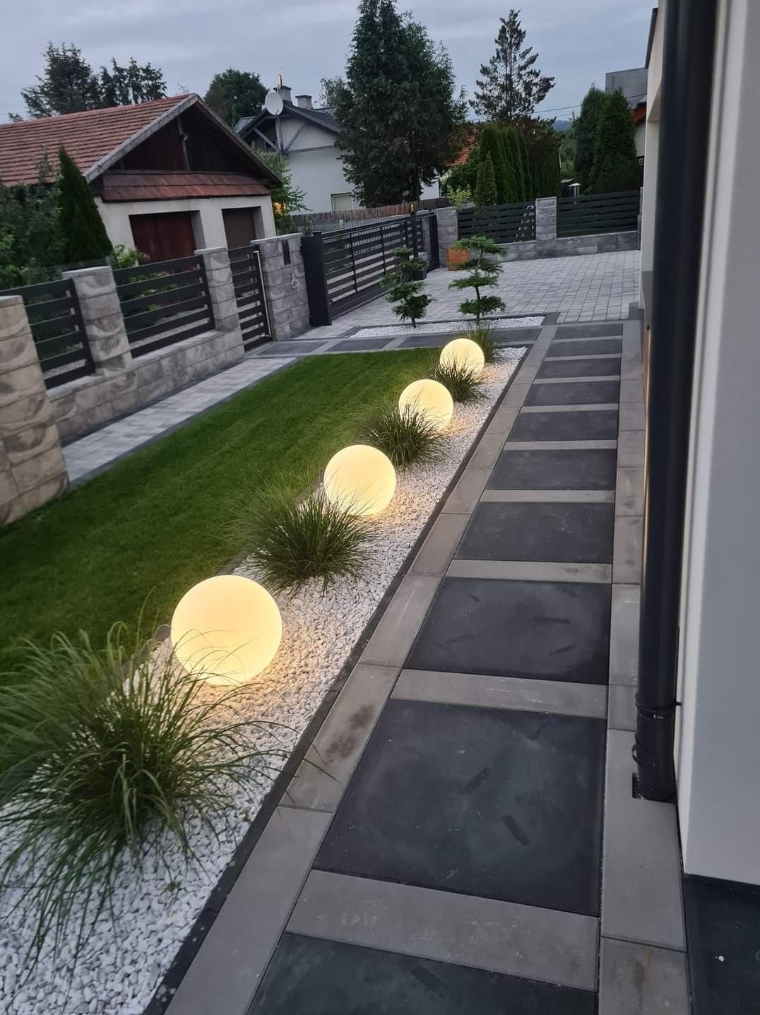 Oświetlony ogród z kulistymi lampami wzdłuż ścieżki z kostki, trawa i ozdobne kamienie. Nowoczesny design ogrodu.