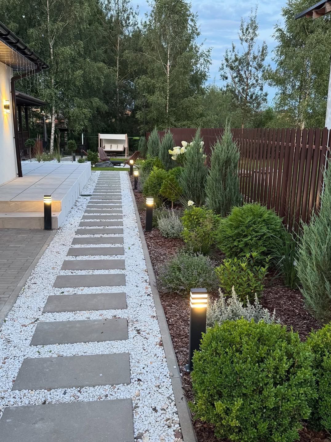 Ogród z ścieżką z płyt, białym żwirem, oświetleniem i ozdobnymi roślinami wzdłuż brązowego ogrodzenia. W tle huśtawka.