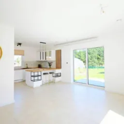 Jasne wnętrze z aneksem kuchennym, beżowe płytki podłogowe, białe ściany, wyspa kuchenna z drewnianym blatem i czarnymi metalowymi elementami, duże okno przesuwne z widokiem na zieleń.