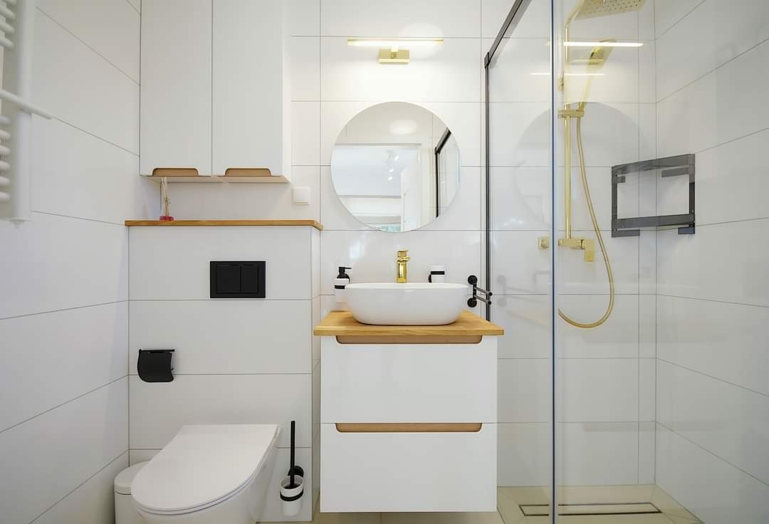 Jasna łazienka z białymi płytkami, okrągłym lustrem, złotą armaturą i kabiną prysznicową typu walk-in z odpływem liniowym.