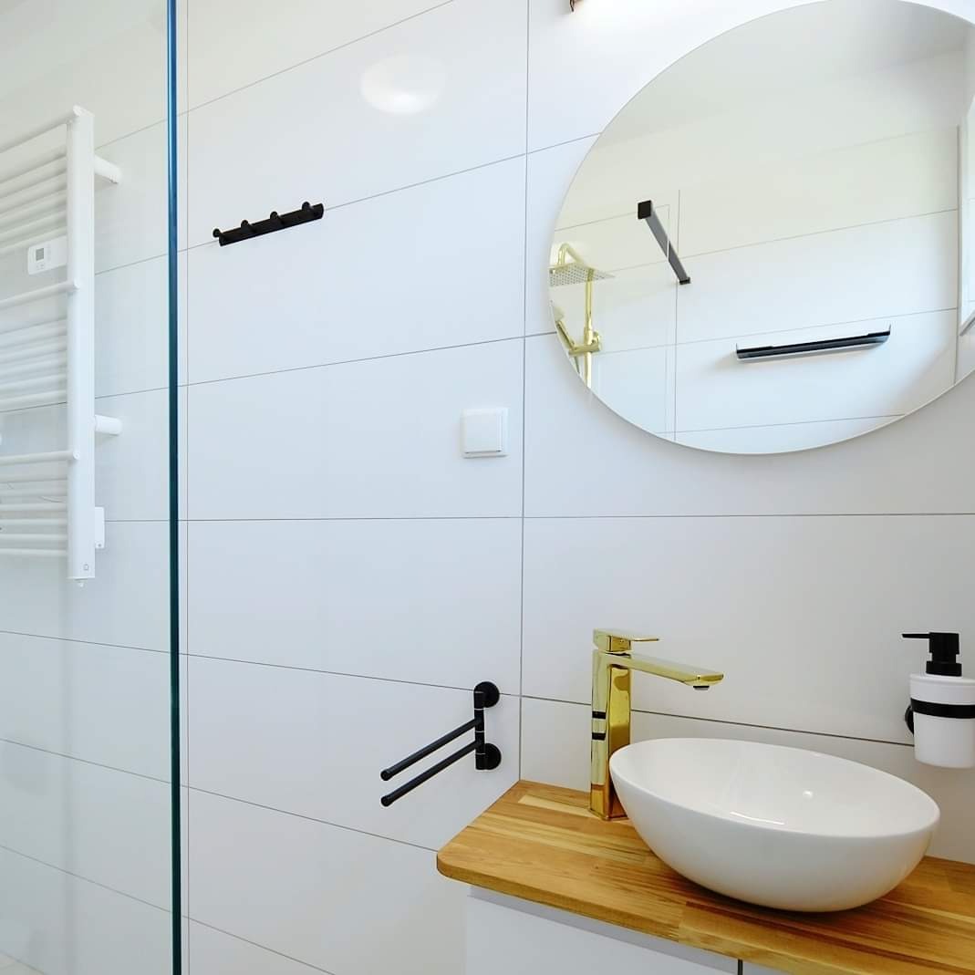 Nowoczesna łazienka z białymi, prostokątnymi płytkami, złotą baterią umywalkową, okrągłym lustrem i drewnianym blatem pod umywalką. Czarny wieszak na ręczniki i dozownik mydła dodają kontrastu.
