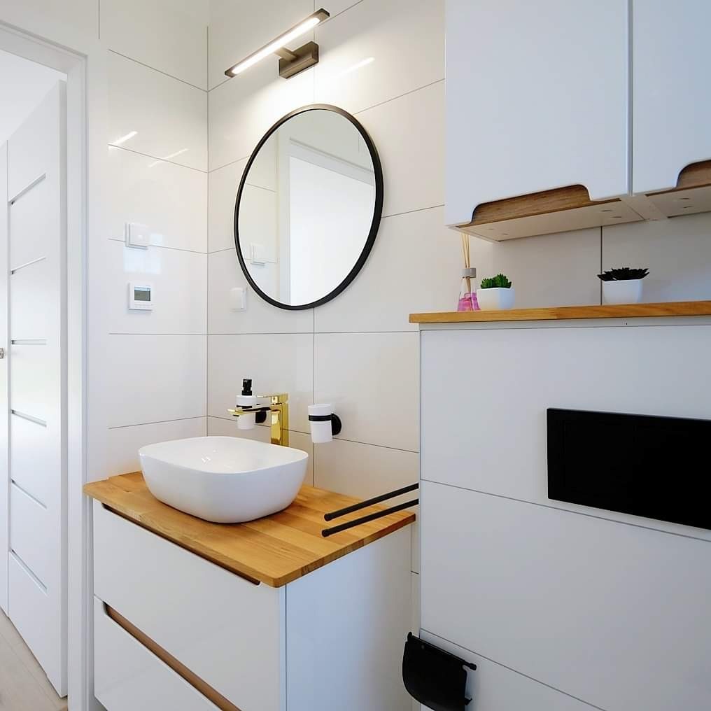 Nowoczesna łazienka z białymi kwadratowymi płytkami, okrągłym lustrem z czarną ramą, blatem z jasnego drewna i białą umywalką nablatową z złotą armaturą.