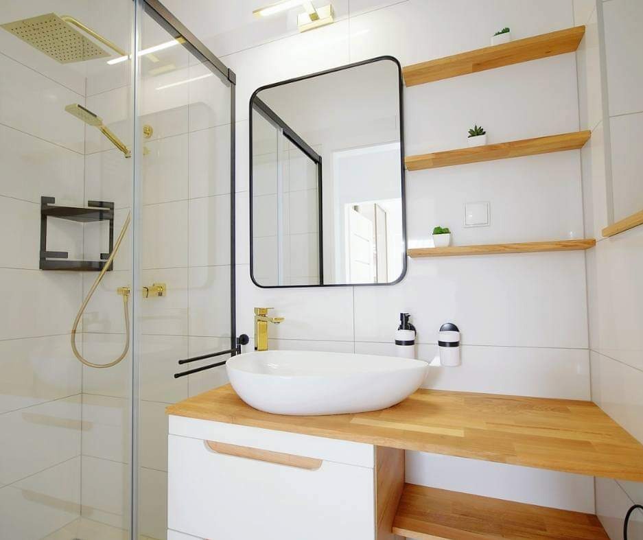 Nowoczesna łazienka z kabiną prysznicową w złotej armaturze, owalną umywalką nablatową i drewnianymi półkami z sukulentami na białych płytkach.