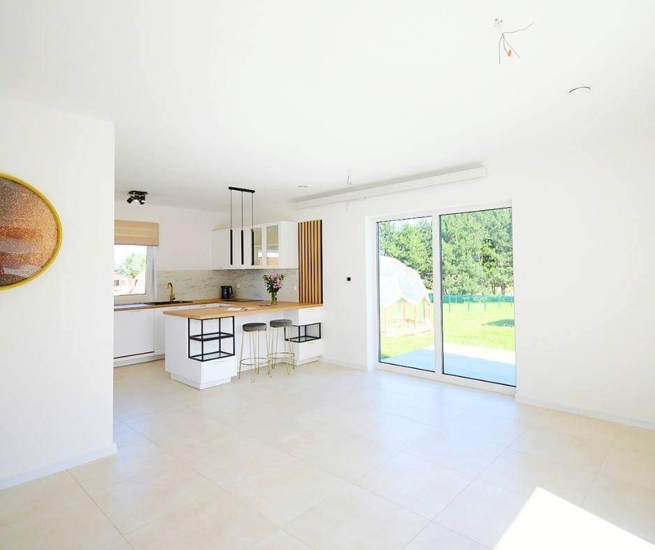 Jasne wnętrze z aneksem kuchennym, beżowe płytki podłogowe, białe ściany, wyspa kuchenna z drewnianym blatem i czarnymi metalowymi elementami, duże okno przesuwne z widokiem na zieleń.