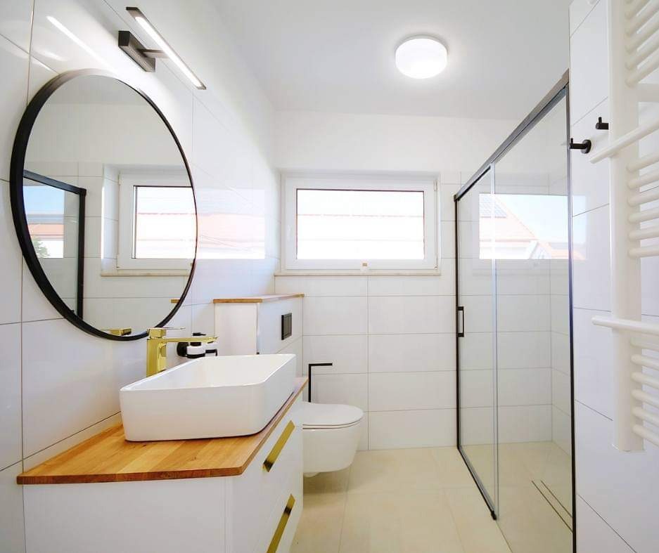 Nowoczesna łazienka w bieli z akcentami drewna i złota: prostokątna umywalka nablatowa, okrągłe lustro w czarnej ramie, kabina prysznicowa z czarnymi profilami, grzejnik drabinkowy i płytki...