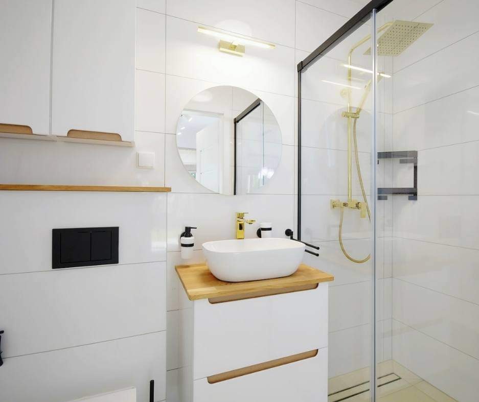 Nowoczesna łazienka z białymi płytkami, okrągłym lustrem, złotą armaturą i kabiną prysznicową typu walk-in z czarnymi elementami.