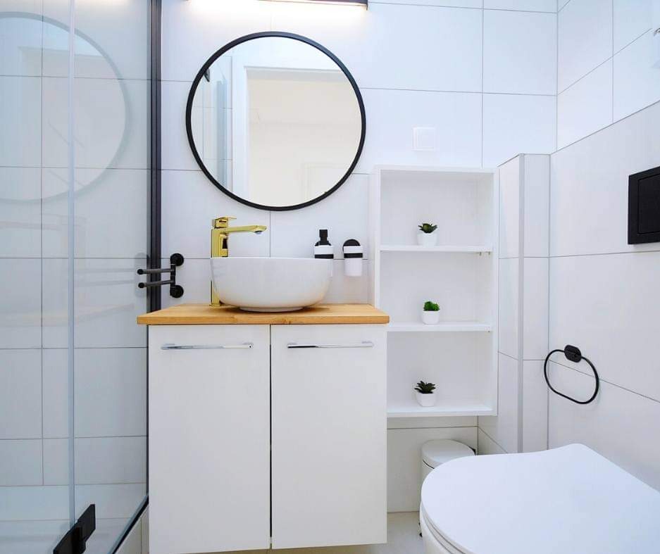 Nowoczesna łazienka w bieli z drewnianym blatem, okrągłym lustrem w czarnej ramie i złotą armaturą, akcenty roślinne na półkach.