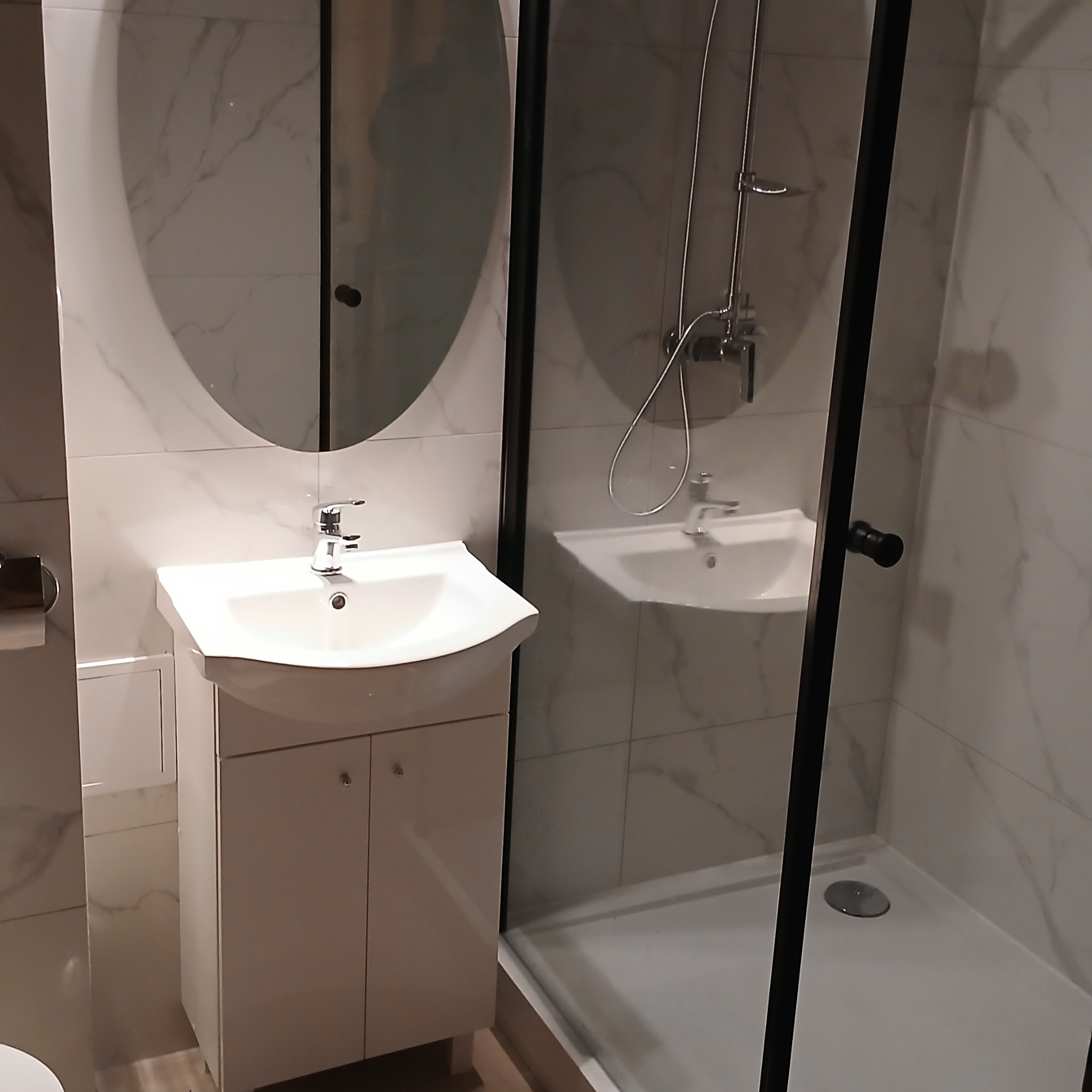Nowoczesna łazienka z umywalką na szafce i kabiną prysznicową typu walk-in, wykończona płytkami imitującymi marmur, z owalnym lustrem bez ramy.